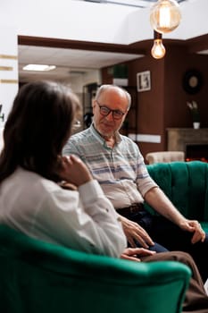 Retired senior husband and wife seated on comfortable sofas in lounge area planning their vacation activities. Happy elderly couple discussing holiday itinerary at exclusive hotel reception.