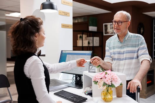 Detailed image shows caucasian retired senior man receiving access card from female receptionist at registration counter. Elderly male tourist giving room key to employee at front desk in hotel lobby.