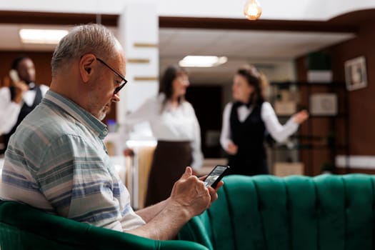 Retired senior man enjoys online connectivity in hotel lobby, using mobile device for communication and relaxation. Elderly male traveler relaxing with smartphone in luxury lounge area.