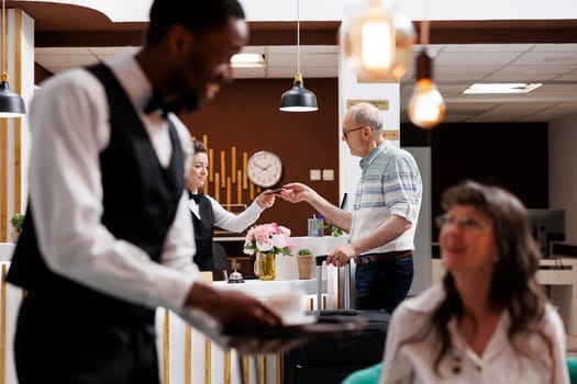 Caucasian receptionist helps retired old man with hotel check-in verifying identity and retrieving reservation details. Female concierge holds and checks passport of male tourist. Focus on background.