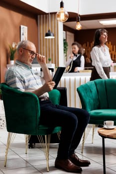 Caucasian elderly male client in hotel lounge area sitting on comfortable sofa with digital tablet. Retired senior man surfing the net for fun vacation activities on his smart device in a nice resort.