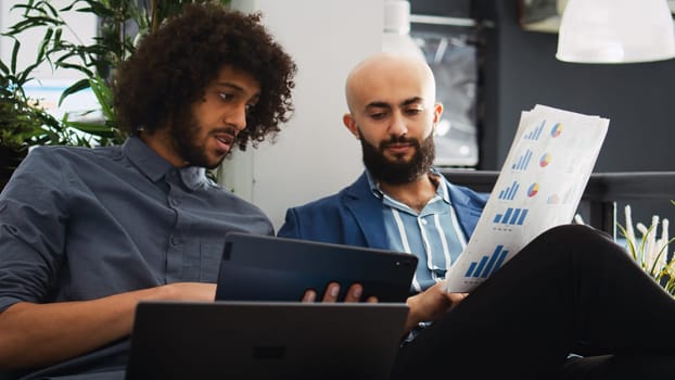 Startup team verify sales report with archived documents and information on laptop, comparing data to create briefing presentation. Coworkers discussing small business development strategy.