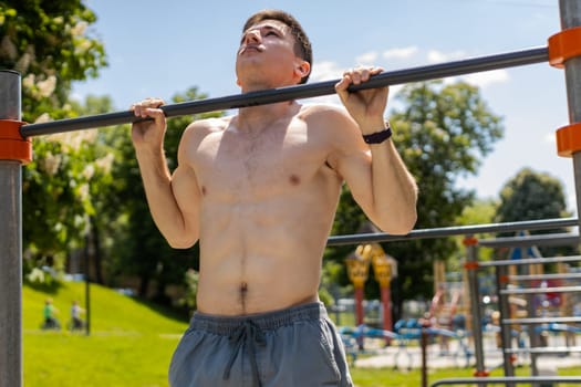 Athletic caucasian topless muscular man doing pull-ups exercises on horizontal bar. Guy pumping up back muscles on summer playground. Sport health fitness routine workout. Bodybuilding motivation