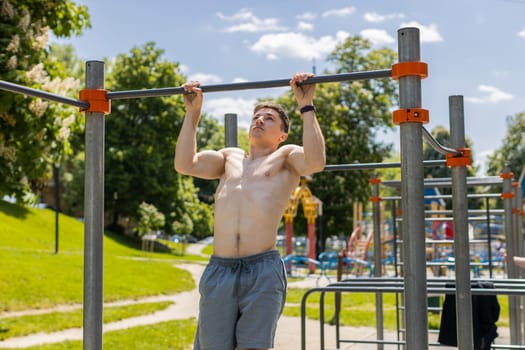 Athletic caucasian topless muscular man doing pull-ups exercises on horizontal bar. Guy pumping up back muscles on summer playground. Sport health fitness routine workout. Bodybuilding motivation