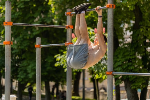 Athletic topless muscular man training abs doing abdominal crunches pull-ups exercises on horizontal bar. Young shirtless guy on playground. Sports, health, fitness routine workout strength gymnastics