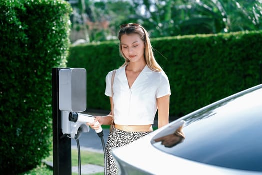 Young woman travel with EV electric car charging in green sustainable city outdoor garden in summer. Urban sustainability lifestyle by green clean rechargeable energy of electric BEV vehicle innards