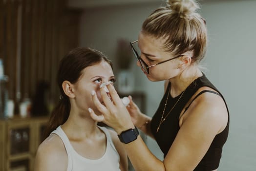 One young handsome Caucasian makeup artist applies a moisturizing medicinal liquid to the lower eyelid under the eye of a girl sitting in a chair early in the morning in a beauty salon, close-up side view.Step by step.