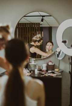 One young handsome Caucasian makeup artist applies protective cream to the face of a girl sitting in a chair, reflected in a round wall mirror early in the morning in a beauty salon, close-up side view. Step by step.