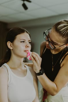 One young handsome Caucasian makeup artist applies foundation with a pink sponge to the chin of a girl sitting in a chair early in the morning in a beauty salon, close-up side view. Step by step.