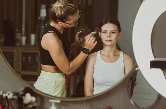 One young handsome Caucasian makeup artist applies foundation powder to a girl's face with a fluffy brush, reflected in a round wall mirror early in the morning in a beauty salon, close-up side view. Step by step.