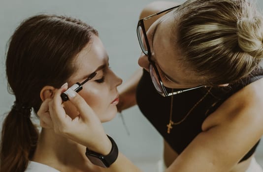 One young handsome Caucasian makeup artist paints with a pencil the eyebrows of a girl sitting sideways in a chair early in the morning in a beauty salon, close-up top view with depth of field. Step by step.