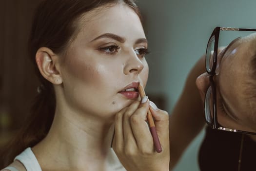 One young handsome Caucasian makeup artist paints with a pencil the lips of a girl sitting sideways in a chair early in the morning in a beauty salon, close-up side view with depth of field. Step by step.