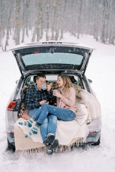 Wife tells something to her husband while he drinks coffee while sitting on blankets in the trunk of a car. High quality photo