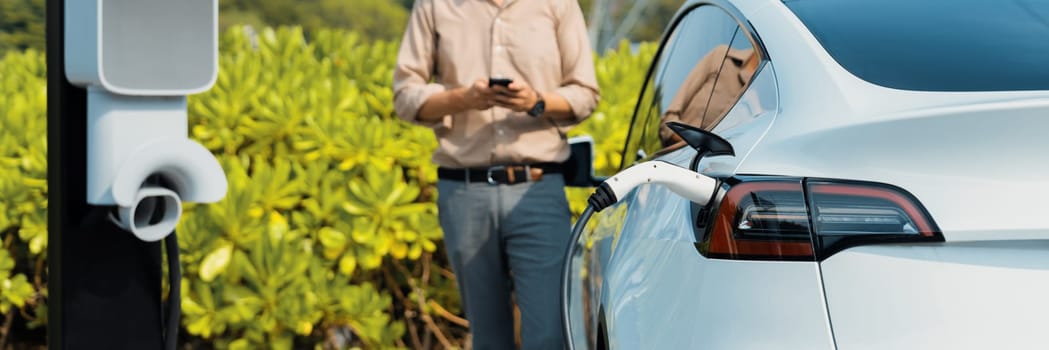 Young man use smartphone to pay for electricity at public EV car charging station green city park. Modern environmental and sustainable urban lifestyle with EV vehicle. Panorama Expedient