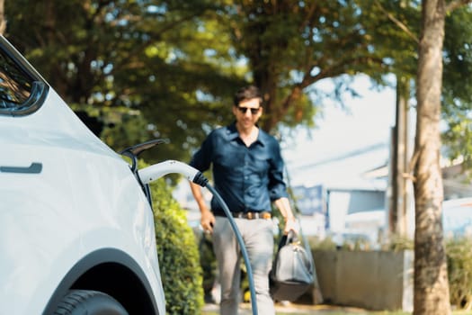 Young man recharge electric car's battery from charging station in outdoor green city park in springtime. Rechargeable EV car for sustainable environmental friendly urban travel lifestyle. Expedient