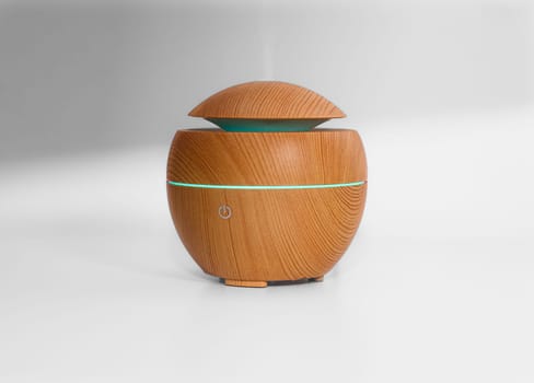 The wellness benefits of aromatherapy are clearly felt in this minimalist portrait of a scented oil electric ultrasonic diffuser and humidifier as it mists on a white blurred background. The fresh wood finish, natural green lamp light and calm, purifying steam vapor suggest a spa like zen atmosphere.