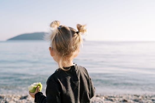 Little girl with an apple stands on the shore and looks at the sea. Back view. High quality photo