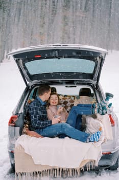 Man kisses a woman on the forehead while sitting in the car trunk with mugs of coffee under snowfall in the forest. High quality photo