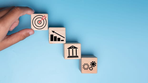 Human hands hold wooden blocks with target icons and business symbols on blue background, business goals and objectives concept, business competition.