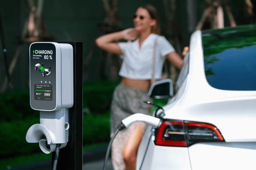 Young woman travel with EV electric car charging in green sustainable city outdoor garden in summer. Urban sustainability lifestyle by green clean rechargeable energy of electric BEV vehicle innards