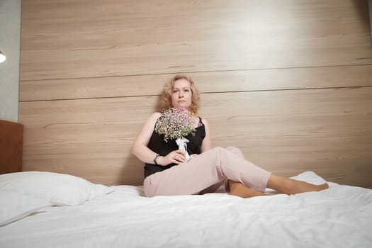 Pretty blonde woman have fine on her bed in bedroom with a bouquet of small flowers. Adult lady enjoying her morning and wake up. The bride before the wedding. A girl on a festive day with gift