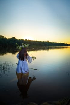 Adult mature brunette woman in a white dress, sundress and a wreath of flowers in summer in water of river or lake in evening at sunset. Celebration of the Slavic pagan holiday of Ivan Kupala