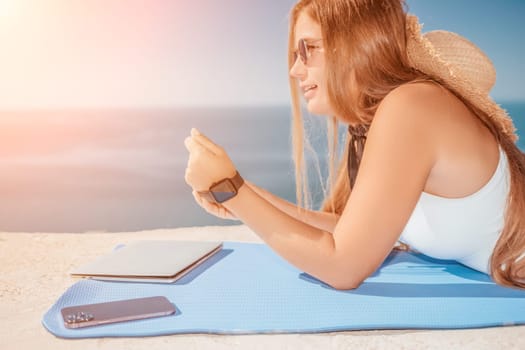 Successful business woman in yellow hat working on laptop by the sea. Pretty lady typing on computer at summer day outdoors. Freelance, travel and holidays concept.