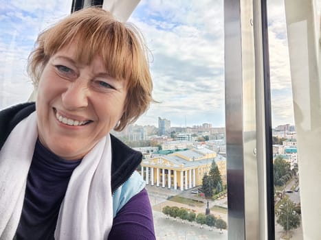 Relaxed mature caucasian female making selfie outside near big Ferris wheel. Positive senior woman against city street. Adult middle agged girl Blogger taking photo for internet