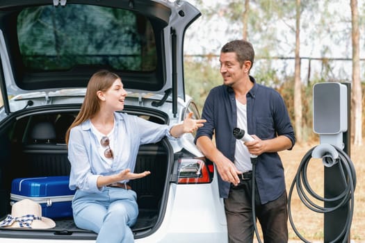 Lovely young couple recharging battery for electric car while relaxing and sitting on the trunk during road trip travel EV car in autumnal forest. Eco friendly travel on vacation during autumn. Exalt