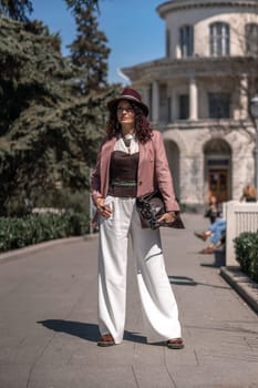 Woman park city. Stylish woman in a hat walks in a park in the city. Dressed in white corset trousers and a pink jacket with a bag in her hands