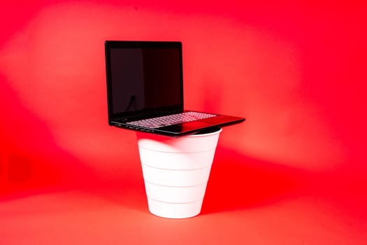 Black laptop in a trash bin on red background isolated