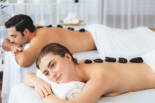 Hot stone massage at spa salon in luxury resort with day light serenity ambient, blissful couple customer enjoying spa basalt stone massage glide over body with soothing warmth. Quiescent