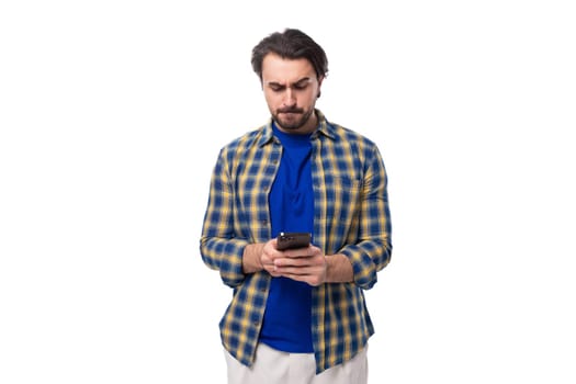 young caucasian brunette with a beard is dressed in a blue plaid shirt and t-shirt watching a video on the phone on a white background.