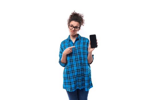 cute young pretty woman with curled hair in a blue shirt shows the screen of a smartphone with a mockup.