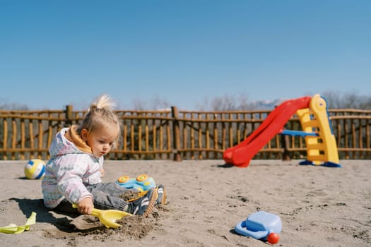 Little girl sits on the playground and digs the sand with a toy shovel. High quality photo