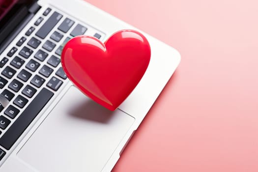 Close up laptop keyboard with a red heart over a pink table with copy space. Online dating concept.