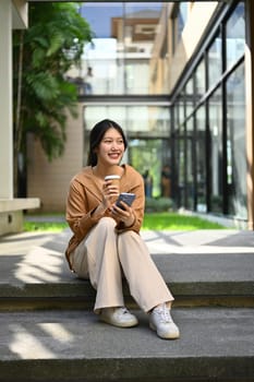 Portrait of young businesswoman with take away coffee cup using mobile phone while sitting near office building.