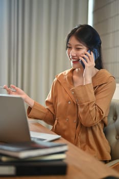 Smiling Asian female employee having pleasant phone conversation and using laptop at workplace.