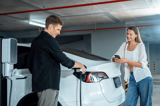 Young couple travel with EV electric car to shopping center parking lot charging in downtown city showing urban sustainability lifestyle by green clean rechargeable energy of electric vehicle innards