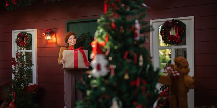 Happy woman wearing Santa hat holding of gift box. Positive emotional Santa girl. with a beautifully decorated Christmas tree serving as the background. festive Xmas concept.