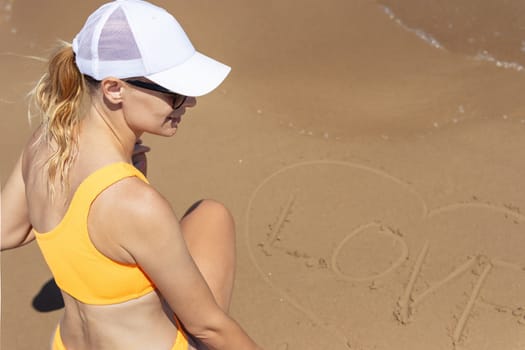 a girl of European appearance in an orange swimsuit and a white cap, sits on the seashore on the sand, draws, there is a place for an inscription. High quality photo