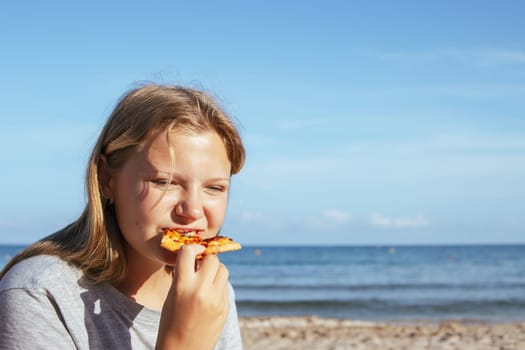 teenage girl of European appearance eats a slice of pizza on the seashore. High quality photo