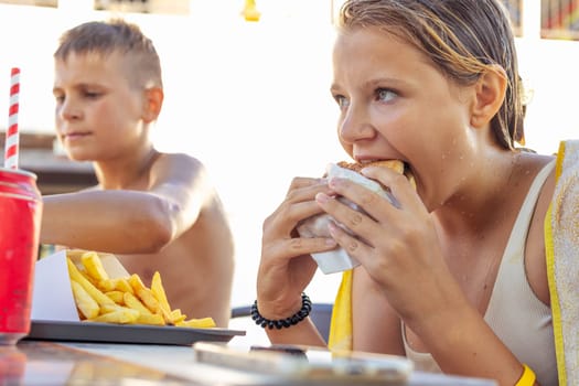 teenagers boy and girl eat burgers and fries on the beach, summer holiday concept. Focus on girl, unhealthy food, children eat unhealthy food, fast food. High quality photo