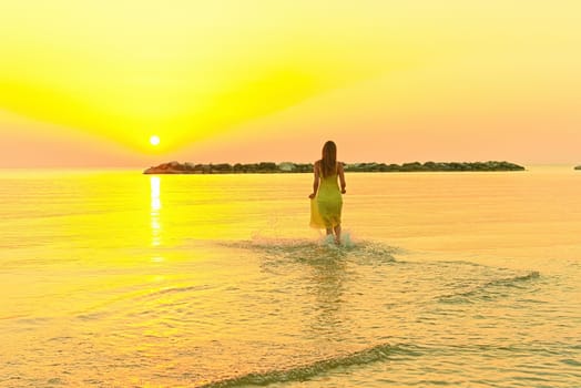 A young woman in a bright yellow dress meets the sunrise, joyfully runs through the sea water