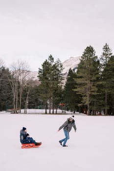 Dad is carrying mom with a child on a sled across a snowy plain looking back. Side view. High quality photo