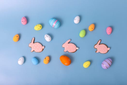 decorative multicolored eggs and three pink gingerbread in the form of rabbit, scattered on a light blue background, top view. Copy space