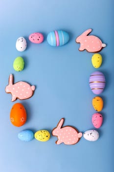 Rectangular frame of multicolored Easter eggs and gingerbread in the form of pink rabbits on a light blue background, top view