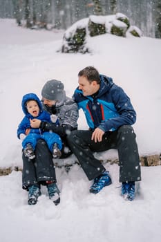 Smiling dad looks at a little girl catching snowflakes with her mouth in her mother arms while sitting on a log in the forest. High quality photo