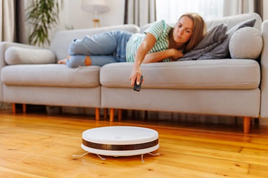 Robot vacuum cleaner cleaning a room while a woman relaxes on the sofa. A woman operates a vacuum with a remote control