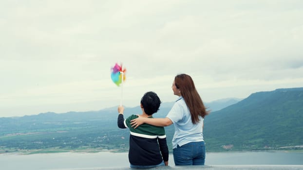 A progressive woman and her son are on vacation, enjoying the natural beauty of a lake at the bottom of a hill while the boy carries a toy windmill.